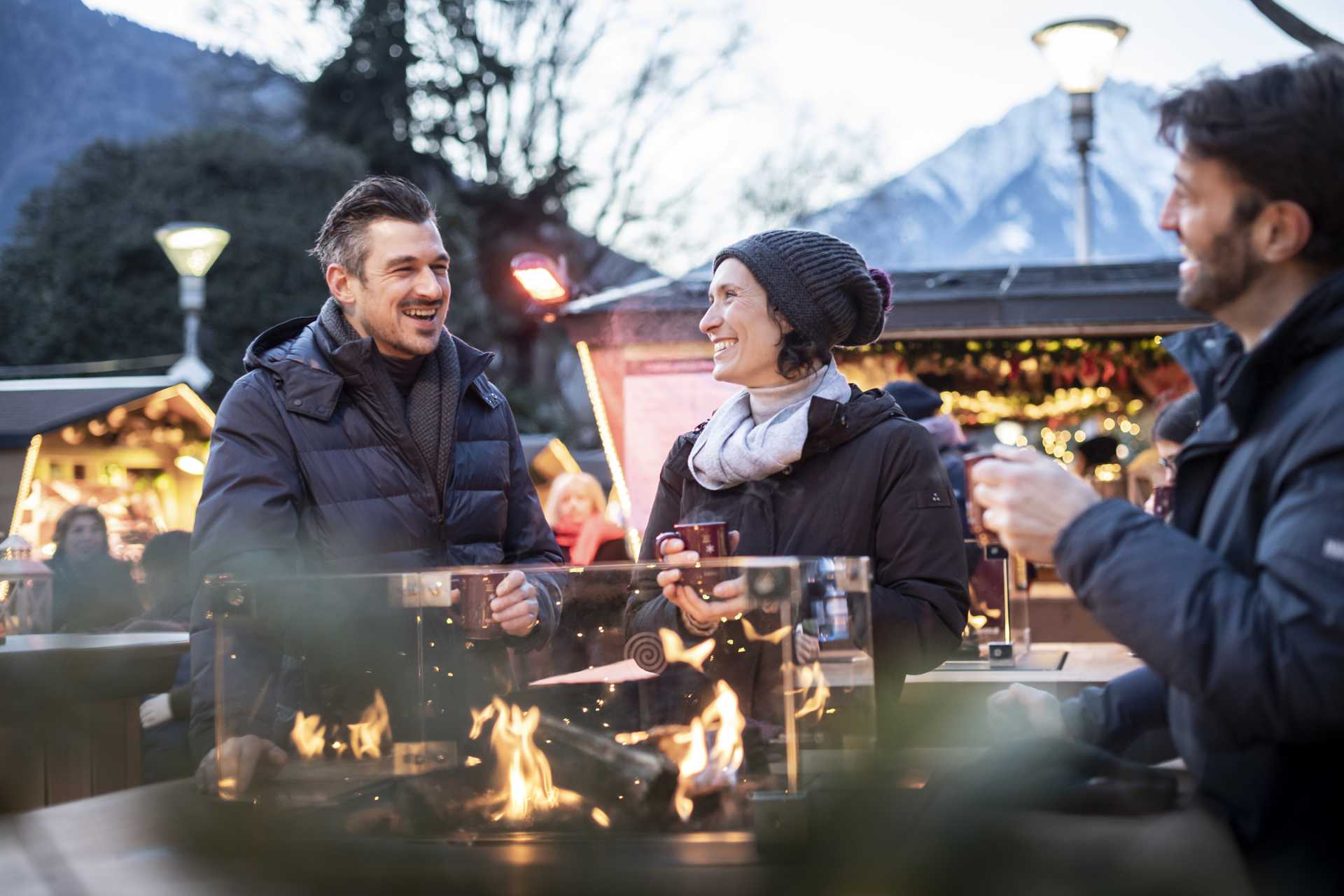 3 Personen die Glühwein trinken und um ein wärmendes Feuer stehen
